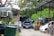 An outdoor area filled with cars, some stacked on top of each other, indicating a car junkyard or repair facility. Vehicles appear in various states of disassembly, with an open hood on the black car in the middle. A forklift is visible near the right, surrounded by green foliage and trees.