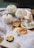 A cozy Halloween scene featuring white pumpkins, spider decorations, and cookies in the shape of skulls and a jack-o'-lantern. The setup includes a burning candle placed on a saucer with cobweb-like material scattered across an open book.