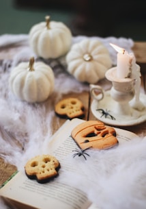 A cozy Halloween scene featuring white pumpkins, spider decorations, and cookies in the shape of skulls and a jack-o'-lantern. The setup includes a burning candle placed on a saucer with cobweb-like material scattered across an open book.