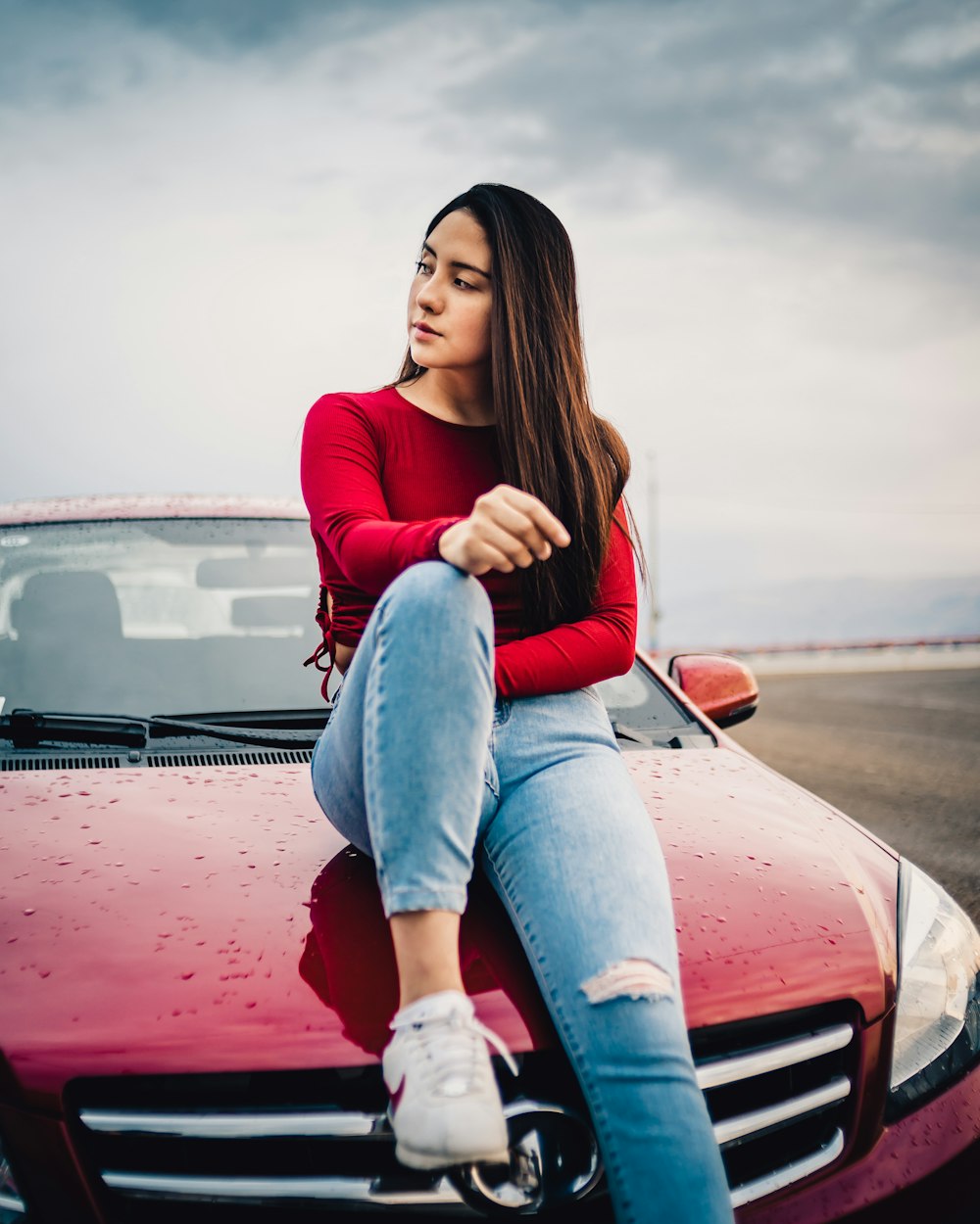 woman in red long sleeve shirt and blue denim jeans sitting on red car hood during
