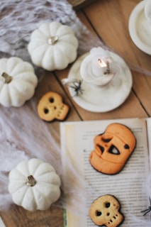 A cozy Halloween-themed setup featuring small white pumpkins, skull-shaped cookies, a lit candle, and faux spider webs on a wooden surface. An open book adds to the seasonal atmosphere, while a tiny black spider enhances the Halloween motif.
