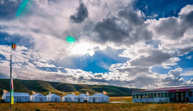 A group of white tents is arranged in a row on a grassy field, with a backdrop of rolling hills. A tall flagpole with multiple colorful flags stands at one end. The sky is expansive, filled with dramatic clouds illuminated by the sun peeking through, creating a vibrant play of light and shadow.