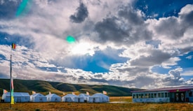 A group of white tents is arranged in a row on a grassy field, with a backdrop of rolling hills. A tall flagpole with multiple colorful flags stands at one end. The sky is expansive, filled with dramatic clouds illuminated by the sun peeking through, creating a vibrant play of light and shadow.