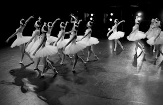 A group of ballet dancers wearing white tutus and headpieces are performing on a dimly lit stage. The dancers are arranged in a coordinated formation, with some extending their arms delicately while others pivot gracefully. Bright stage lights are visible in the background, creating a contrast with the shadows on the floor.