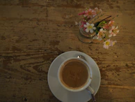 A coffee cup on a rustic wooden table next to a notebook and a vase of fresh flowers.