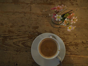 A cozy coffee cup resting beside a small vase of fresh flowers on a wooden table.