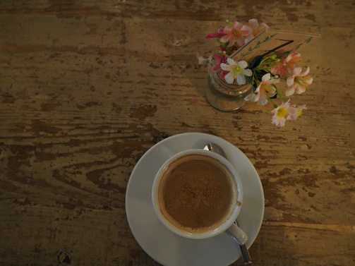 A cozy coffee cup resting beside a small vase of fresh flowers on a wooden table.