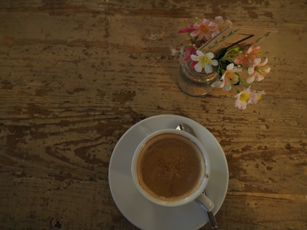 A steaming cup of coffee on a rustic wooden table beside a small vase of fresh flowers.