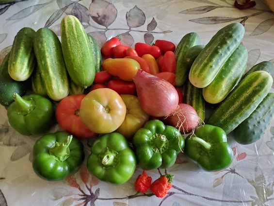 An assortment of fresh vegetables including cucumbers, red tomatoes, green bell peppers, and onions are arranged together on a patterned surface. The vegetables display a variety of shapes and vibrant colors, indicating freshness and abundance.