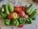An assortment of fresh vegetables including cucumbers, red tomatoes, green bell peppers, and onions are arranged together on a patterned surface. The vegetables display a variety of shapes and vibrant colors, indicating freshness and abundance.