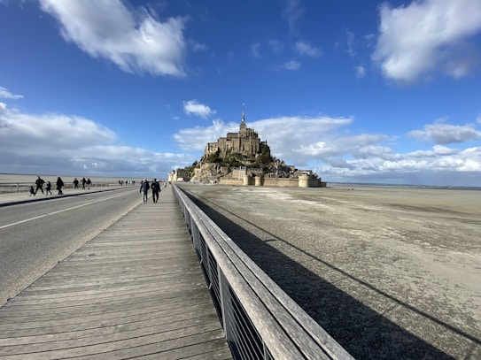A stunning view of Mont Saint-Michel, an ancient island fortress surrounded by sand and a wide causeway leading to it. The sky is blue with scattered clouds, and several people are walking along the path.