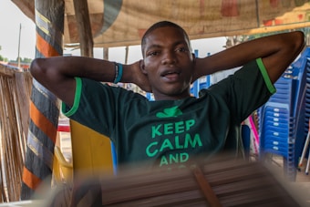 A person wearing a green shirt with the words 'Keep Calm' is sitting with their hands resting on the back of their head. They appear relaxed, sitting under a shaded area with striped orange and black decorations and stacked blue plastic chairs in the background.