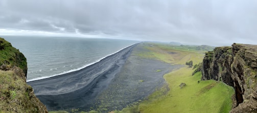 A vast coastline with a dramatic black sand beach stretches alongside the ocean. The landscape features steep cliffs covered in patches of lush green grass, contrasting sharply with the dark sand. The sky is overcast, casting a soft light over the scene, adding to the natural beauty and serene atmosphere.