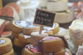 A variety of cheeses are displayed in a market setting. In the foreground, there is a small round cheese with a black chalkboard sign inserted into it, reading 'Évora Pequeno D.O.P. 2,50€'. Other cheeses can be seen in the background, each with labels and various shapes and sizes.