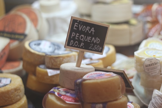 A variety of cheeses are displayed in a market setting. In the foreground, there is a small round cheese with a black chalkboard sign inserted into it, reading '&Eacute;vora Pequeno D.O.P. 2,50&euro;'. Other cheeses can be seen in the background, each with labels and various shapes and sizes.