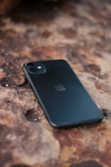 A smartphone with a black case is lying on a textured, rusty-colored surface. The phone is positioned diagonally, and the surface appears to be rough and uneven.