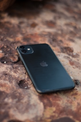 A smartphone with a black case is lying on a textured, rusty-colored surface. The phone is positioned diagonally, and the surface appears to be rough and uneven.