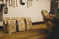 A series of brown paper bags labeled 'Family Farm' with sizes such as small, medium, and large printed on them are placed on a wooden counter. A person's hands are seen arranging one of the bags. In the background, various knives and a power outlet are attached to the wall.