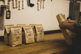 A series of brown paper bags labeled 'Family Farm' with sizes such as small, medium, and large printed on them are placed on a wooden counter. A person's hands are seen arranging one of the bags. In the background, various knives and a power outlet are attached to the wall.