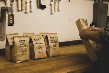 A series of brown paper bags labeled 'Family Farm' with sizes such as small, medium, and large printed on them are placed on a wooden counter. A person's hands are seen arranging one of the bags. In the background, various knives and a power outlet are attached to the wall.