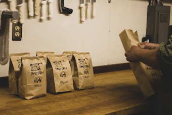 A series of brown paper bags labeled 'Family Farm' with sizes such as small, medium, and large printed on them are placed on a wooden counter. A person's hands are seen arranging one of the bags. In the background, various knives and a power outlet are attached to the wall.