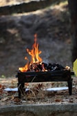 A small barbecue grill sits on the ground with a lively fire burning inside. Orange flames and dark charcoal are visible, with some smoke rising. The grill is surrounded by dry leaves and grass, indicating an outdoor setting with a natural, earthy backdrop.