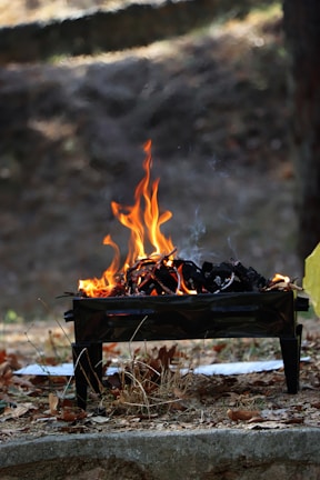 A small barbecue grill sits on the ground with a lively fire burning inside. Orange flames and dark charcoal are visible, with some smoke rising. The grill is surrounded by dry leaves and grass, indicating an outdoor setting with a natural, earthy backdrop.