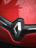 Close-up view of a red car with water droplets covering its surface. The car's logo is prominently displayed, featuring a silver emblem with a black background.
