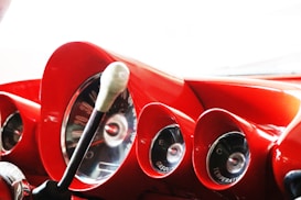 Close-up view of a vintage car dashboard with red accents, featuring circular gauges for speed, gasoline, and temperature. The gear shift lever is prominently displayed in the foreground, adding to the retro aesthetic.