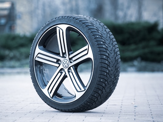 A single car tire with a detailed tread pattern is positioned upright on a brick pavement. The tire features a shiny alloy wheel with a Volkswagen logo in the center. The background is slightly blurred, highlighting the tire as the main focus.
