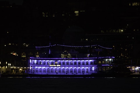 A riverboat with multiple decks is illuminated by blue and white lights, creating a striking visual against the backdrop of a dark cityscape. The name 'Louisiana Star' is visible on the side of the boat. Strings of smaller lights adorn the upper decks, enhancing the boat's ornate structure.