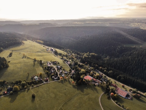 An aerial view of a rural landscape featuring a small village surrounded by lush green fields and dense forests. The village consists of several houses and buildings scattered along winding roads. The scene is bathed in soft lighting with sunlight filtering through clouds, casting long shadows across the fields and trees.