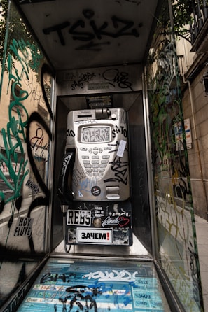 An old phone booth covered in various graffiti and stickers, featuring a metallic payphone in the center. The walls of the booth are transparent and adorned with tags, scribbles, and colorful designs. The scene is set against an urban backdrop with a nearby building visible through the glass.