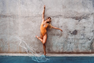 A person in a vibrant orange swimsuit strikes a dynamic pose against a textured, weathered wall. Their outstretched arm and extended leg suggest movement and grace, complemented by water splashing slightly at their feet.