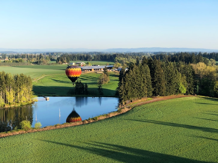 A vibrant hot air balloon floats above a lush green landscape with rolling hills and a clear blue sky. Below, a serene pond reflects the balloon and surrounding trees. Farm buildings are in the distance, bordered by dense forests.