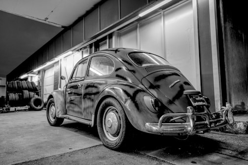 A vintage car is parked inside a garage with industrial lighting. The classic vehicle has a rounded shape, typical of early mid-century models. The garage features rows of tires stacked on the left side.