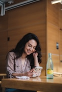 A woman is sitting at a wooden table in a well-lit setting, smiling as she looks at her phone. A bottle of sparkling mineral water is placed on the table alongside a small cloth or face mask.