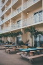 A multi-story building with several balconies featuring white railings and outdoor chairs. Below, there are picnic tables with blue and green accents lined along a patio area, surrounded by potted palm trees and various plants. String lights hang above the tables, adding a cozy and inviting atmosphere.