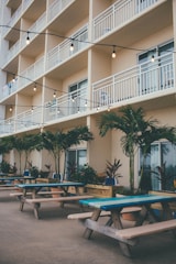 A multi-story building with several balconies featuring white railings and outdoor chairs. Below, there are picnic tables with blue and green accents lined along a patio area, surrounded by potted palm trees and various plants. String lights hang above the tables, adding a cozy and inviting atmosphere.