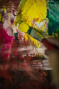 A paintbrush is actively applying vibrant strokes of yellow paint onto a colorful abstract canvas, with surrounding hues of pink, red, green, and blue blending together. The image captures the dynamic motion of painting.