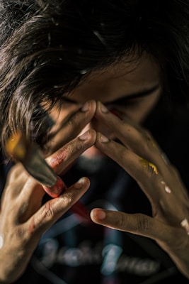 A close-up of a person with their hands near their face, covered in paint. The focus is on the hands, which are holding a paintbrush, suggesting artistic activity. The lighting creates a dramatic shadow effect, emphasizing the texture of the paint on the hands.