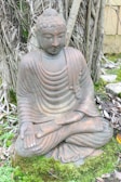 A stone statue of a seated Buddha in a meditative posture surrounded by lush green moss and foliage. The statue is positioned against a backdrop of tree roots, creating a natural, serene setting.