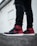 A pair of stylish sneakers with black and red coloring worn with ripped black jeans. The setting appears urban, with a white wall in the background.