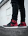 A pair of stylish sneakers with black and red coloring worn with ripped black jeans. The setting appears urban, with a white wall in the background.