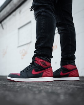 A pair of stylish sneakers with black and red coloring worn with ripped black jeans. The setting appears urban, with a white wall in the background.