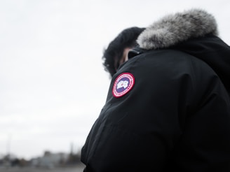A person is wearing a dark parka with a fur-lined hood and a distinct round patch featuring a logo on the sleeve. The background is a cloudy, overcast sky, and the focus is on the jacket.