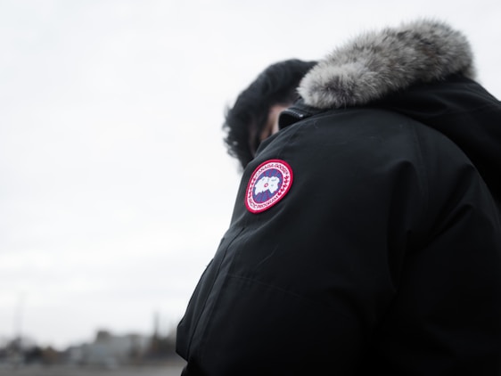 A person is wearing a dark parka with a fur-lined hood and a distinct round patch featuring a logo on the sleeve. The background is a cloudy, overcast sky, and the focus is on the jacket.