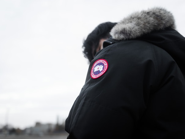 A person is wearing a dark parka with a fur-lined hood and a distinct round patch featuring a logo on the sleeve. The background is a cloudy, overcast sky, and the focus is on the jacket.