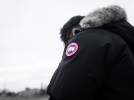 A person is wearing a dark parka with a fur-lined hood and a distinct round patch featuring a logo on the sleeve. The background is a cloudy, overcast sky, and the focus is on the jacket.