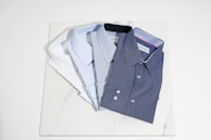 A row of neatly folded shirts in satin, twill, and pinpoint oxford fabrics, arranged by color gradient.